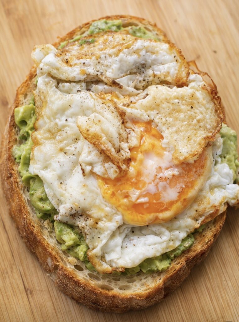 Whole-grain toast topped with mashed avocado and two crispy fried eggs on a wooden cutting board